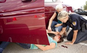 Vehicle accident with driver on ground. Passenger and policewoman checking her injuries. Police vehicle in background.