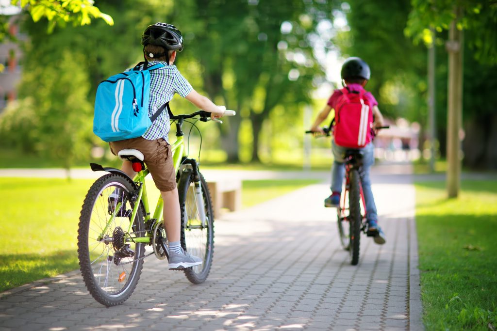 Children,With,Rucksacks,Riding,On,Bikes,In,The,Park,Near