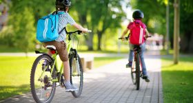 Children,With,Rucksacks,Riding,On,Bikes,In,The,Park,Near